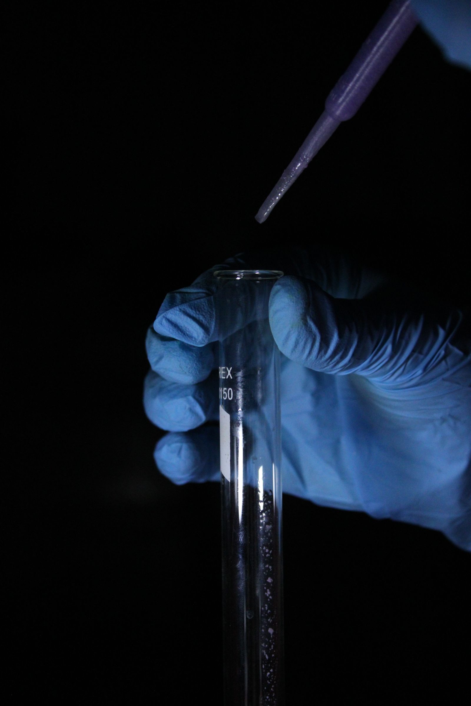 Blue-gloved hand holding a test tube beneath a pipette against a dark background.